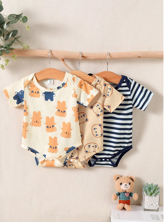 Three baby bodysuits with different patterns hanging on a rack, with a small teddy bear toy and plant in the background.