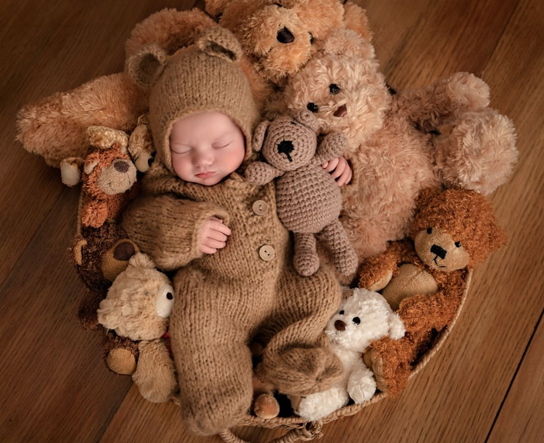 Baby in a knitted brown outfit surrounded by teddy bears on a wooden floor.